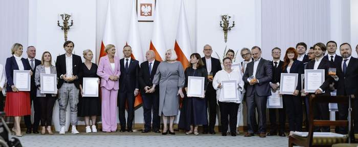 Pokaż zdjęcie: W półokręgu stoi wiele elegancko ubranych osób niektórzy trzymają w rękach oprawione dyplomy. Pośrodku Pan i Pani Prezydent. Za nimi flagi polskie.