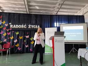 Pokaż zdjęcie: Obok mównicy stoi kobieta blondynka, w jasnej marynarce i ciemnych spodniach, w tle ekran z prezentacją. Na ścianie zrobiono hasło radość życia oraz przyczepiono kolorowe motyle.