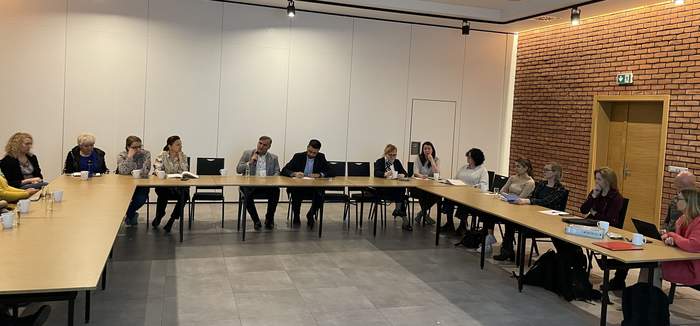 Pokaż zdjęcie: Przy długim drewnianym stole w kształcie litery U siedzą ludzie. W centralnym miejscu siedzi Ksiądz obok niego mężczyzna w jasnoszarej marynarce trzymający mikrofon.