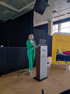 Pokaż zdjęcie: Kobieta, blondynka w zielonym garniturze stoi przy mównicy, zdjęcie podczas konferencji.