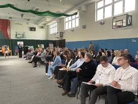 Pokaż zdjęcie: Kilkadziesiąt osób, głównie dorosłych, siedzi w rzędach krzeseł w sali gimnastycznej i uważnie słucha osoby przemawiającej z lewej strony. Uczestnicy są elegancko ubrani, część trzyma notatki lub dokumenty. W tle widoczne są kosze do koszykówki i duże okna wpuszczające światło.