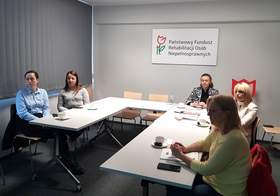 Grupa osób siedzących przy stole konferencyjnym w kształcie liter „U”, na których leżą notesy oraz napoje. Uczestnicy spotkania zwróceni są w kierunku prowadzącego spotkanie. Na prawej ścianie wisi prostokątna tablica z logo w kształcie tulipana i napisem: „Państwowy Fundusz Rehabilitacji Osób Niepełnosprawnych”. 