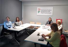 Pokaż zdjęcie: Grupa osób siedzących przy stole konferencyjnym w kształcie liter „U”, na których leżą notesy oraz napoje. Uczestnicy spotkania zwróceni są w kierunku prowadzącego spotkanie. Na prawej ścianie wisi prostokątna tablica z logo w kształcie tulipana i napisem: „Państwowy Fundusz Rehabilitacji Osób Niepełnosprawnych”. 