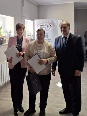 Pokaż zdjęcie: Trzy osoby stojące w holu Warsztatu Terapii Zajęciowej w Gołańczy pozujące do zdjęcia: dwie osoby od lewej trzymają białe teczki z dokumentami. Osoby ubrane są elegancko w garnitury, marynarki oraz eleganckie bluzki. Po lewej stoi kobieta w krótkich brązowych włosach, w różowej marynarce i ciemnych spodniach. Pośrodku stoi kobieta w okularach, w jasnej bluzce z dużym napisem na przodzie i trzyma ciemną torbę. Po prawej stoi mężczyzna w garniturze i krawacie, z krótkimi włosami.