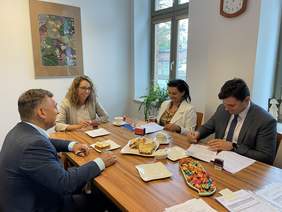 Pokaż zdjęcie: Przy drewnianym stole siedzi dwóch mężczyzn i dwie kobiety. Wszyscy ubrani w marynarki. Na stole leżą dokumenty, pieczątki i filiżanki. Mężczyzna i kobieta podpisują dokumenty uśmiechając się.