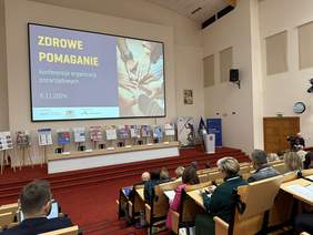 Pokaż zdjęcie: W audytorium na fotelach siedzą uczestnicy spotkania. Przy mównicy stoi kobieta. Obok niej stoją roll-upy organizatorów i plakaty organizacji pozarządowych. Nad nimi wisi duży ekran wyświetlający tytuł konferencji. 