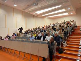 Pokaż zdjęcie: W auli siedzą ludzie. Są to słuchacze konferencji. Wszyscy z zainteresowaniem słuchają wykładów.  