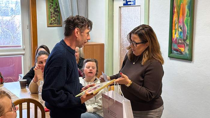 Pokaż zdjęcie: Kobieta wręcza mężczyźnie dyplom w złotej ramie, w tle przy stołach siedzą uczestnicy WTZ.