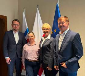 Uczestnicy spotkania w Kołobrzegu pozują do wspólnego zdjęcia na tle flag PFRON, Polski i Unii Europejskiej. Na zdjęciu: Dyrektor Zachodniopomorskiego Oddziału PFRON Bartosz Baszczyński, przedstawicielka Stowarzyszenia „Zamknięty Świat”, Prezydent Kołobrzegu Anna Mieczkowska oraz Starosta Kołobrzeski Mirosław Tessikowski.