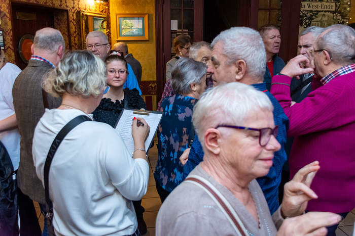 Pokaż zdjęcie: Tłum osób w trakcie rozmów i rejestracji przy wejściu. Na pierwszym planie kobieta w białej bluzce trzymająca kartkę z listą, a w tle osoby w różnym wieku w luźnej atmosferze świątecznej.