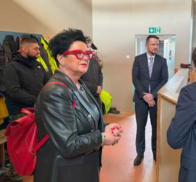 Pokaż zdjęcie: Prezes Borowiec ubrana w czarna sukienkę i skórzaną , czarną kurtkę, ma charakterystyczne czerwone okulary, podziwia budynek MZAZ.