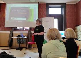 Pokaż zdjęcie: Kobieta w czarnej bluzce z napisem OWiT przemawia do zgromadzonych osób za nią na ekranie wyświetla się prezentacja.