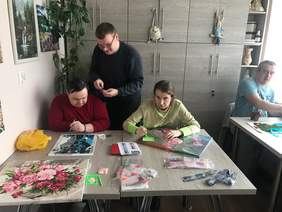 Pokaż zdjęcie: Po lewej stronie przy stole siedzi mężczyzna z kobietą na stole rozłożone są obrazy i materiały do ich tworzenia nad nimi stoi pochylony ubrany na czarno mężczyzna, po prawej stronie przy stoliku siedzi mężczyzna w niebieskiej koszulce.