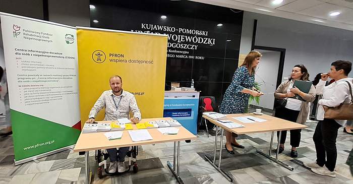 Punkt informacyjny Kujawsko-Pomorskiego Oddziału PFRON i punktu CIDON. Przy stole siedzi pracownik zamiejscowego punktu CIDOn PFRONm a przed nim leżą materiały informacyjne.  Za nim stoją banery  CIDON i Dostępność, a obok widoczne jest kolejne stanowiski, pzry któym stpją trzy kobiety.