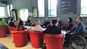 Pokaż zdjęcie: Uczestnicy posiedzenia Rady siedzą na fotelach, przy stole, słuchając przekazu Pani Dyrektor Gierej, w tle widoczny ekran z prezentacją