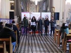 Pokaż zdjęcie: miejsce w Kościele pod wezwaniem Św. Józefa Oblubieńca NMP w Ozorkowie po środku stoją osoby trzymają mikrofony ubrani są w czapki Mikołajowe, śpiewają dla publiczności 