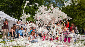 Pokaż zdjęcie: Zdjęcie przedstawia grupę osób dorosłych i dzieci podrzucających rolki z papierem toaletowym w parku.