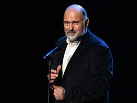 Pokaż zdjęcie: Mężczyzna z siwą brodą i krótkimi włosami stoi przy mikrofonie, ubrany w ciemną marynarkę i jasną koszulę. Patrzy w stronę widowni z lekkim uśmiechem.