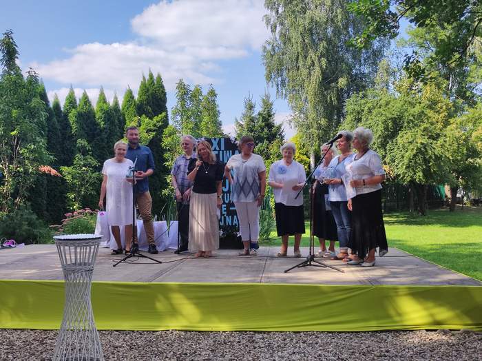Pokaż zdjęcie: Zdjęcie w ogrodzie, na zbudowanej scenie stoją laureaci festiwalu pieśni maryjnej, łącznie 8 osób, dwóch mężczyzn i 6 kobiet.