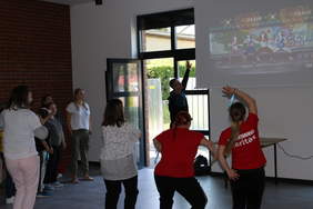 Pokaż zdjęcie: Duża sala, szara kafelkowa podłoga, jedna ze ścian jest wyłożona czerwoną cegłą.  Przez duże szklane wejściowe drzwi wpada dużo słońca. Na środku sali ćwiczy grupa osób wpatrzonych ekran, na którym wyświetlane są taneczne zadania. Stoj