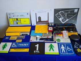 Pokaż zdjęcie: Na stoliku znajdują się gadżetu ułatwiające osobom niewidomym funkcjonowanie w przestrzeni publicznej. Między innymi tabliczki z napisem w alfabecie braille'a, zegarek oraz karty do gry.