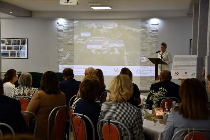 Pokaż zdjęcie: Na ścianie sali konferencyjnej wyświetlana jest prezentacja, na której widoczny jest pałac z lotu ptaka. W sali siedzi grupa kilkunastu osób, które przysłuchują się wystąpieniu kobiety przemawiającej do mikrofonu.