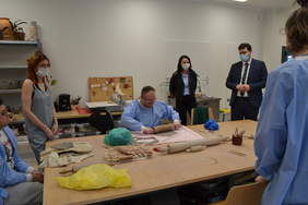 Pokaż zdjęcie: Warsztaty ceramiczne odbywają się przy stole. Jest dwóch uczestników i dwóch opiekunów. Jeden z uczestników wałkuje glinę. Na stole są wałki z różnymi wytłoczonymi wzorami, które odwzorowują się na cieście