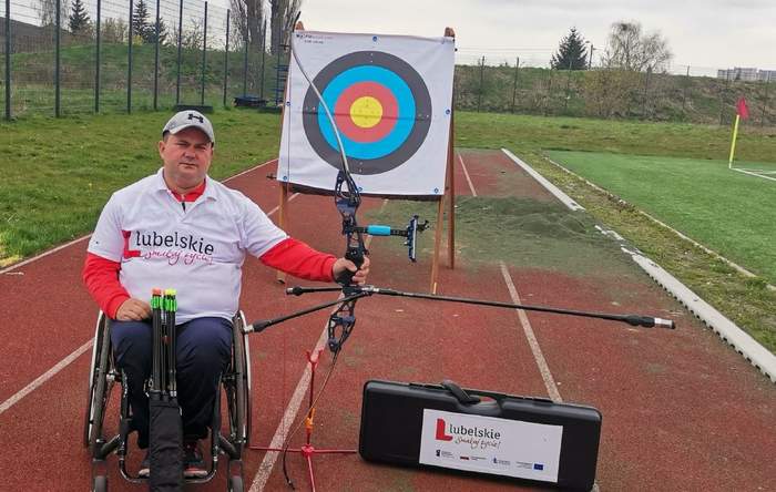 Pokaż zdjęcie: Mężczyzna na wózku inwalidzkim, rękę lewa ma oparta na łuku, za plecami ma  tarczę strzelniczą