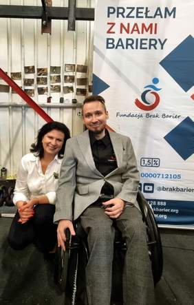 Pokaż zdjęcie: Mężczyzna na wózku inwalidzkim i kobieta siedzą obok siebie. Uśmiechają się. Kobieta po lewej stronie ma ciemne włosy do ramion, białą bluzkę, ciemne spodnie. Mężczyzna w szarej marynarce, ciemnej koszuli i spodniach. Za nim w tle pionowy banner z napisem Przełam z nami bariery, Fundacja brak barier.