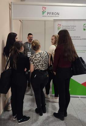 Pokaż zdjęcie: Stoisko targowe z logiem PFRON, mężczyzna w okularach w szarej bluzie udziela informacji stojącym przy stoisku owiedzającym
