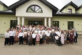 Pokaż zdjęcie: Grupa ludzi pozujących do wspólnego zdjęcia przed wejściem do budynku. Wśród obecnych są uczestnicy WTZ, pracownicy, osoby z niepełnosprawnościami, goście i przedstawiciele lokalnej społeczności.