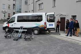 Pokaż zdjęcie: dwa wózki inwalidzkie oraz winda załadowcza, na tle białego samochodu marki Ford Transit, którego stanowią wyposażenie, z tyłu samochodu widoczny jest wysuwany podest, samochód opisany www.mops-znin.pl