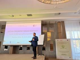 Pokaż zdjęcie: Mężczyzna w garniturze stoi przed ekranem z tekstem "OBSZAR CHRONIONY OBSZAR DOSTĘPNY Model dostępnego parku przyrodniczego Ruciane-Nida, 29-30.XI.2023". Mężczyzna trzyma mikrofon i prezentuje prezentację . Po prawej znajduje się i roll-up 