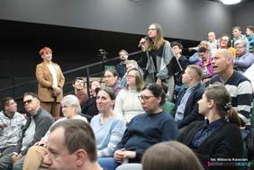 Pokaż zdjęcie: Zdjęcie auli, na krzesłach siedzą uczestnicy konferencji, uczestnicy WTZ, w tle stoi kobieta w beżowym garniturze, z jednego z krzeseł wstała młoda dziewczyna, która zadaje pytanie do mikrofonu