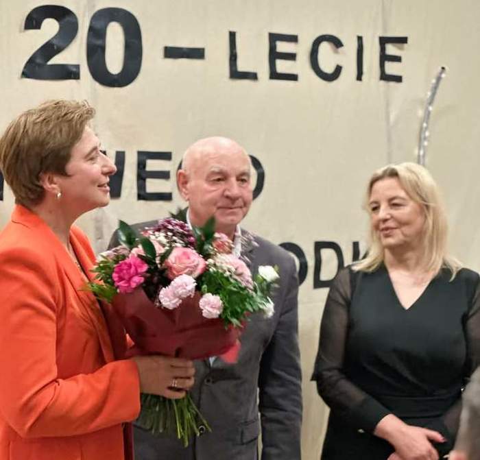 Pokaż zdjęcie: Na zdjęciu trzy osoby, jedna z kobiet trzyma kwiaty, pośrodku kobiet stoi mężczyzna w garniturze.