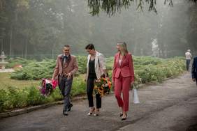 Pokaż zdjęcie: Drogą idą trzy osoby, mężczyzna oraz dwie kobiety. Kobieta i mężczyzna trzymają w ręku duże bukiety kolorowych kwiatów.