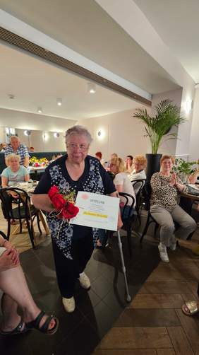 Pokaż zdjęcie: Starsza pani podtrzymująca sie kulą trzyma w rękach dyplom i kwiaty. W tle przy stolikach siedzą starsze osoby. 