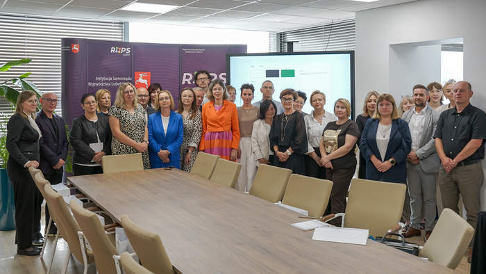 Pokaż zdjęcie: W sali konferencyjnej do pamiątkowego zdjęcia pozuje grupa osób, uczestników spotkania.