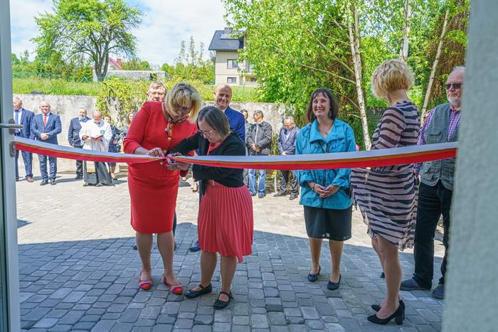 Pokaż zdjęcie: Dyrektor Marta Mordarska oraz Uczestniczka WTZ symbolicznie przecinają wstęgę podczas otwarcia siedziby. W tle goście zaproszeni na uroczystość