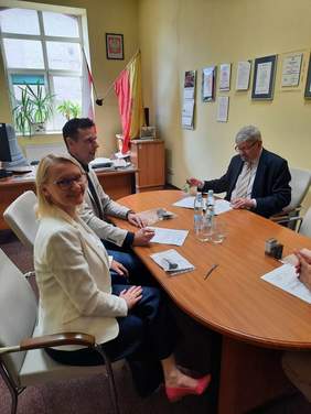 Pokaż zdjęcie: Przy owalnym stole siedzą trzy osoby: kobieta i dwóch mężczyzn. Na stole leżą dokumenty, pieczątki, długopisy, szklanki i woda. Kobieta patrzy w obiektyw aparatu i szeroko się uśmiecha. 