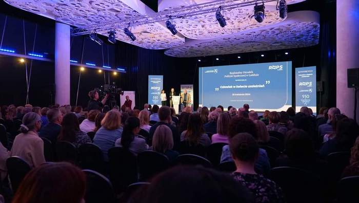 Pokaż zdjęcie: Zdjęcie podczas konferencji zrobione z tyłu sali w kierunku sceny, na dużym ekranie wyświetlana jest prezentacja, po lewej stronie sceny stoi mównica.