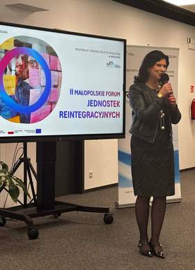 Elegancko ubrana kobieta przemawia do mikrofonu. W tle ekran z napisem: II Małopolskie Forum Jednostek Reintegracyjnych w Krakowie