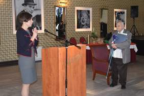 Pokaż zdjęcie: W sali restauracyjnej elegancko ubrana kobieta stoi przed mównicą, mając w dłoniach mikrofon. Po drugiej stronie możemy dostrzec starszego mężczyznę, który trzyma w rękach kulę ortopedyczną oraz teczki z gratulacjami z okazji jubileuszu. W tle widoczne jest zarówno lustro, jak i wielkoformatowe portrety znanych aktorów, które zostały zawieszone na ścianie sali.