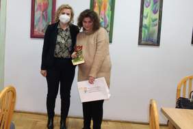 Pokaż zdjęcie: Dwie kobiety stoją przed na sali . Jedna z nich trzyma w  lewej dłoni białą torbę z czerwono- zielonym kwiatkiem i napisem Państwowy Fundusz Rehabilitacji Osób Niepełnosprawnych, w prawej statuetkę  przestawiającą czerwono-zielonego kwiatka 