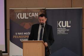 Pokaż zdjęcie: Przy mównicy stoi mężczyzna w średnim wieku ubrany w ciemny garnitur