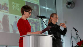 Pokaż zdjęcie: Kobieta w czerwonej sukience i czarnych okularach przemawia przy mównicy. Trzyma w ręku kartkę papieru. Po prawej stronie stoi kobieta w czarnej bluzce i okularach, tłumacz języka migowego. Za nimi duży ekran z kolorową grafiką.