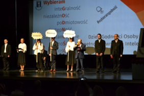 Pokaż zdjęcie: Scena, na której stoi ośmiu artystów ubranych na biało czarno lub w garnitury. Trzy osoby trzymają tabliczki w kształcie chmurki z hasłami: #dostępność, #niezależność, #podmiotowość.