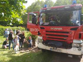 Pokaż zdjęcie: Chłopiec siedzi w wozie strażackim obok strażaka. Ma na głowie hełm strażacki. Przy drzwiach kolejka mam z dziećmi, które chcą wejść do wozu. 