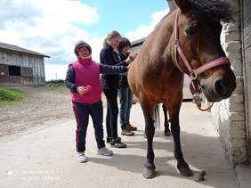 Pokaż zdjęcie: Dwie dziewczyny czyszczą konia wspólnie z terapeutką. Jedna z dziewczyn niewysoka,ubrana w rożowy bezrękawnik, druga wyższa ubrana w czarny sweter 