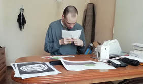 Pokaż zdjęcie: Przy stole siedzi mężczyzna. Jest pochylony i wycina coś nożyczkami. Na stole leżą różne kartki i rysunki. 